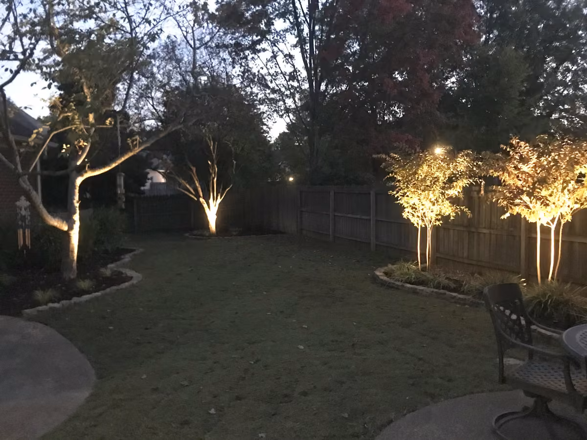 Low-voltage uplights on the trees along a backyard fence at dusk, patio table in the foreground