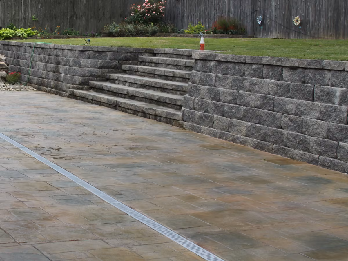 A stone block retaining wall with steps up to the lawn, above a stamped concrete drive