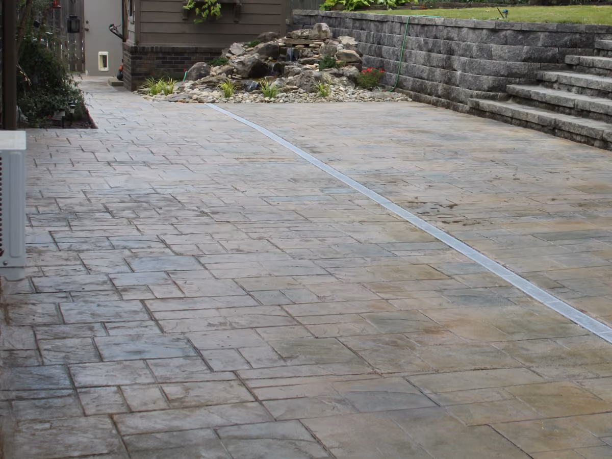 A stamped concrete drive with a channel drain set into it, stone retaining wall behind